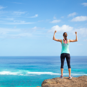 A woman standing on a rock raising her arms in victory in front of the ocean