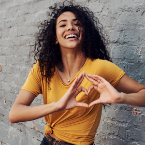 A woman standing against a white brink wall making a heart symbol with her hands