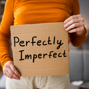 A woman in an orange long sleeved shirt holding up a cardboard sign that reads "Perfectly Imperfect".