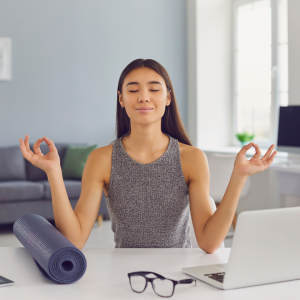 A woman sitting at her work desk raising her hands in a meditative state with her eyes closed