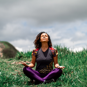 A woman sitting in an open field in a meditative sitting position with her eyes closed and head to the sky