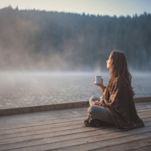 A woman wrapped in a blanket sitting on a dock with a cup of coffee overlooking a lake