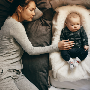 a woman laying down sleep reaching to her baby in the bassinet next to hear