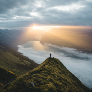 A woman on standing on top of a tall mountain seeing the sunrise peak through the clouds