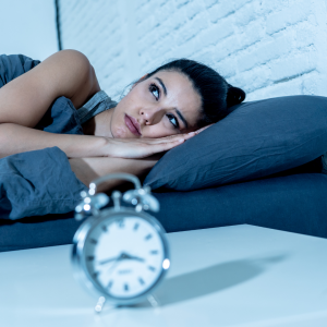 A woman laying down restless on the bed with a clock placed in front of her