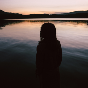 A woman standing on a dark as the sun goes down staring out to the lake